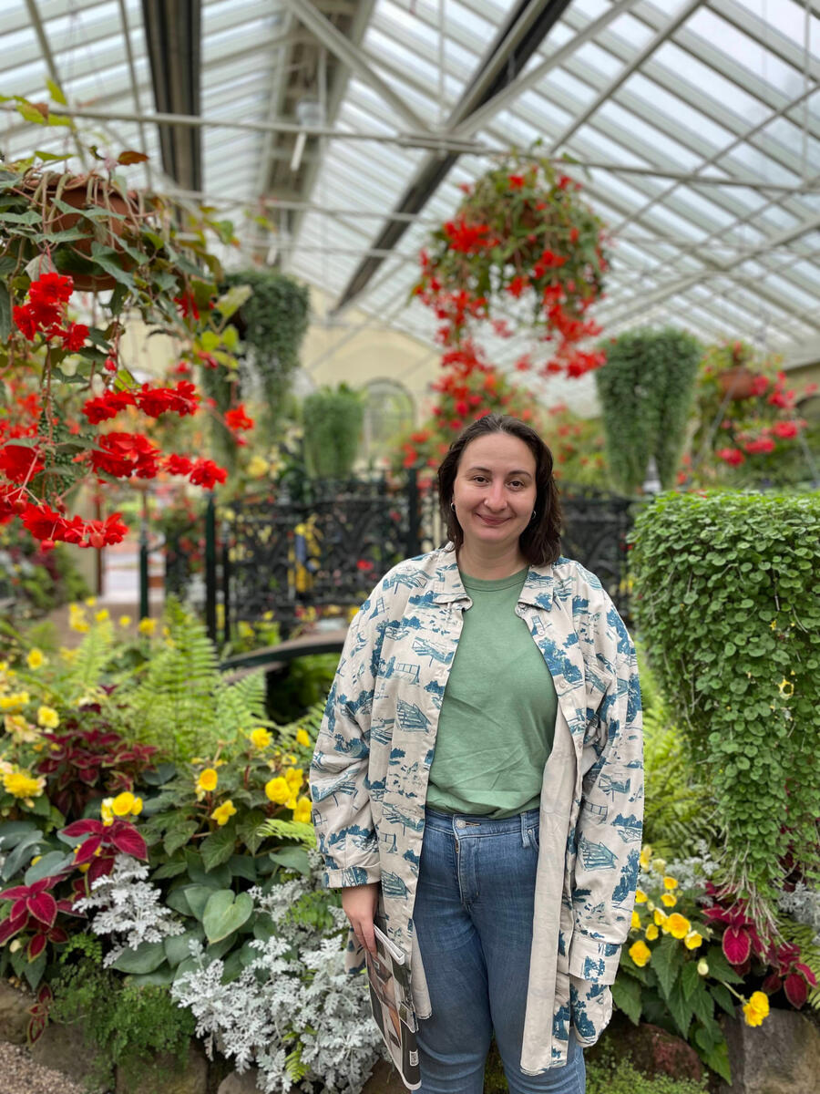 A picture of a woman (Amelia) with brown hair and white skin standing in a greenhouse full of colourful flowers.
