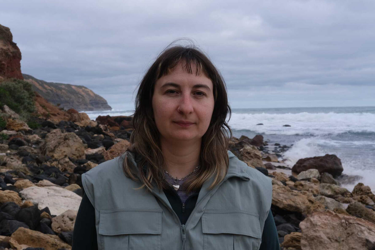 A picture of a woman (Amelia) with brown hair and white skin standing in front of the ocean and rocks.